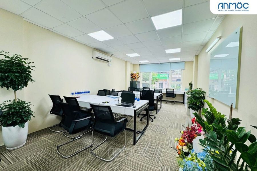 Không gian phòng làm việc với bàn dài màu trắng, nhiều ghế tựa lưng lưới đen, và cây xanh trang trí dọc theo tường