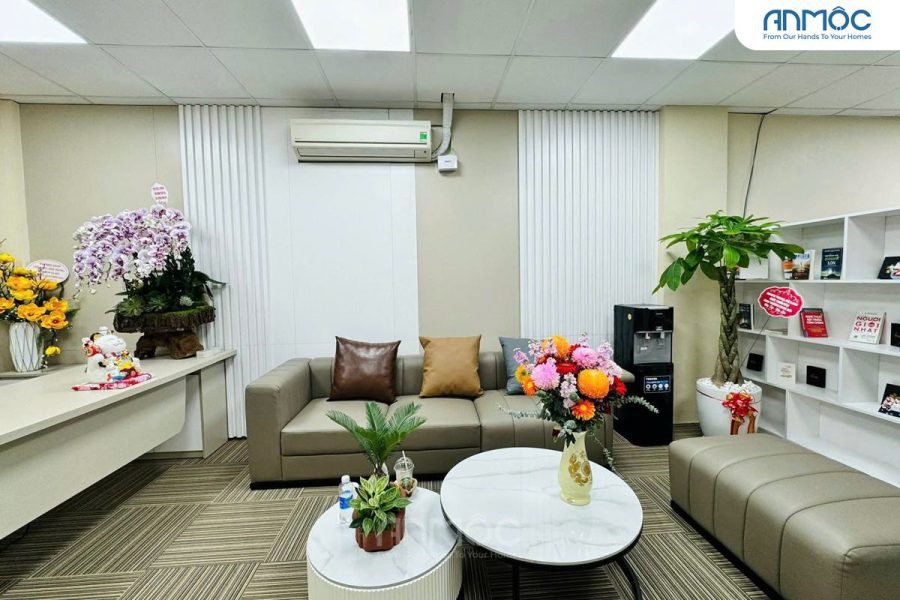 Bàn làm việc lễ tân với bình hoa tươi, chậu cây nhỏ và tượng mèo thần tài may mắn, cạnh khu vực sofa chờ bọc da màu nâu