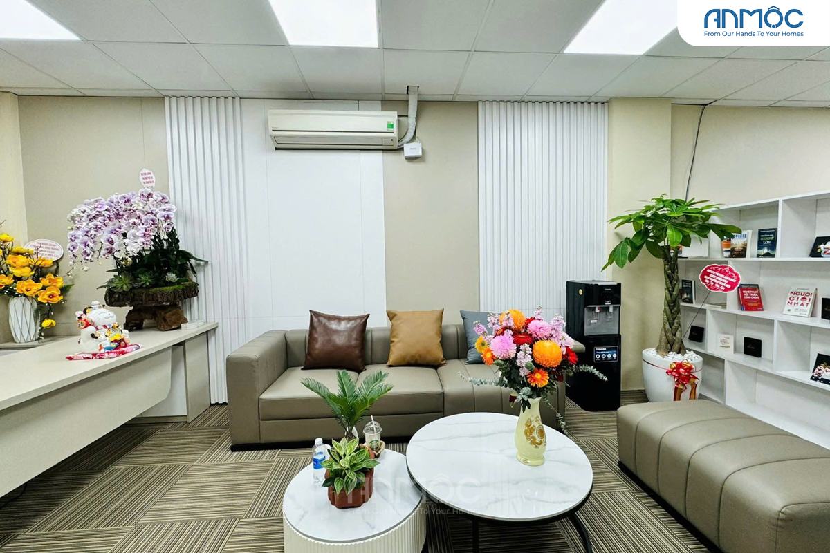 Bàn làm việc lễ tân với bình hoa tươi, chậu cây nhỏ và tượng mèo thần tài may mắn, cạnh khu vực sofa chờ bọc da màu nâu